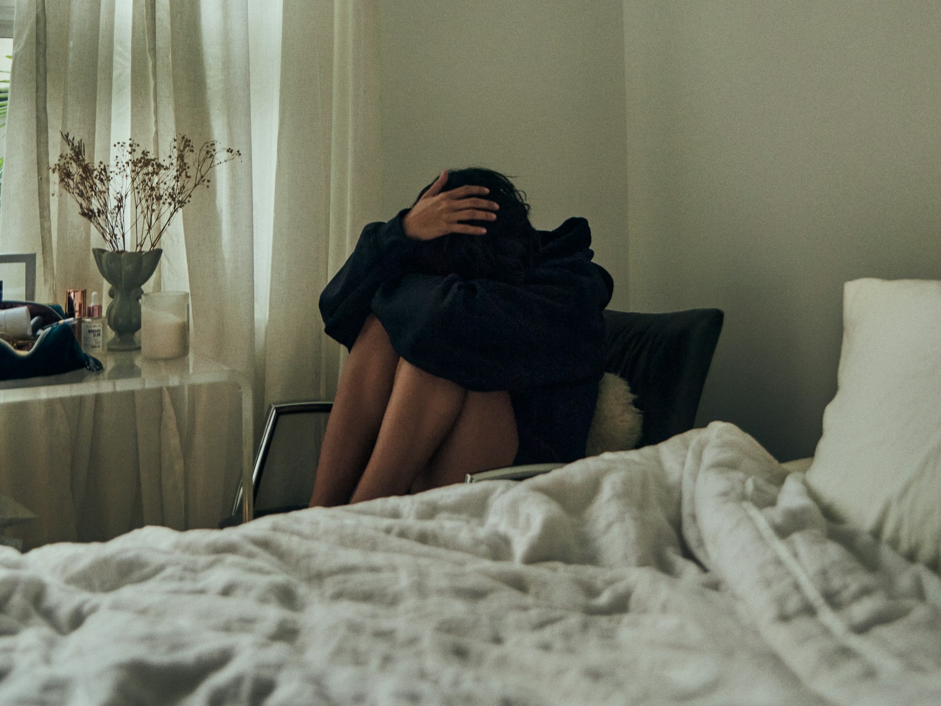 Person sitting on a chair by a bed with knees folded up to their chest, with their face burrowed into their lap.